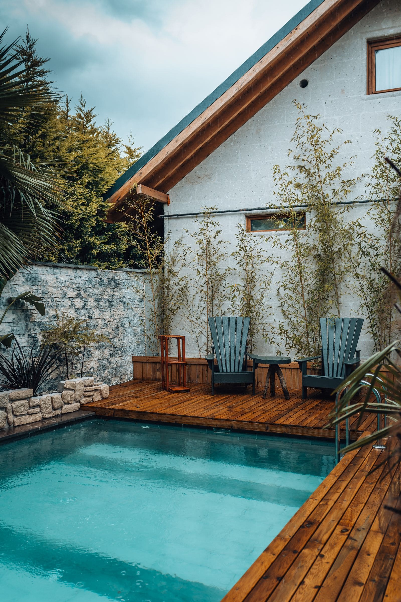Backyard pool beside a white house, with a wooden deck and two outdoor chairs